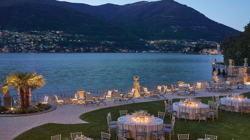 An elegant waterfront celebration setup on Lake Como, featuring candlelit tables and breathtaking mountain views at dusk. Celebrate your next luxury event with bespoke curation by Nadia Forte Travel & Events.