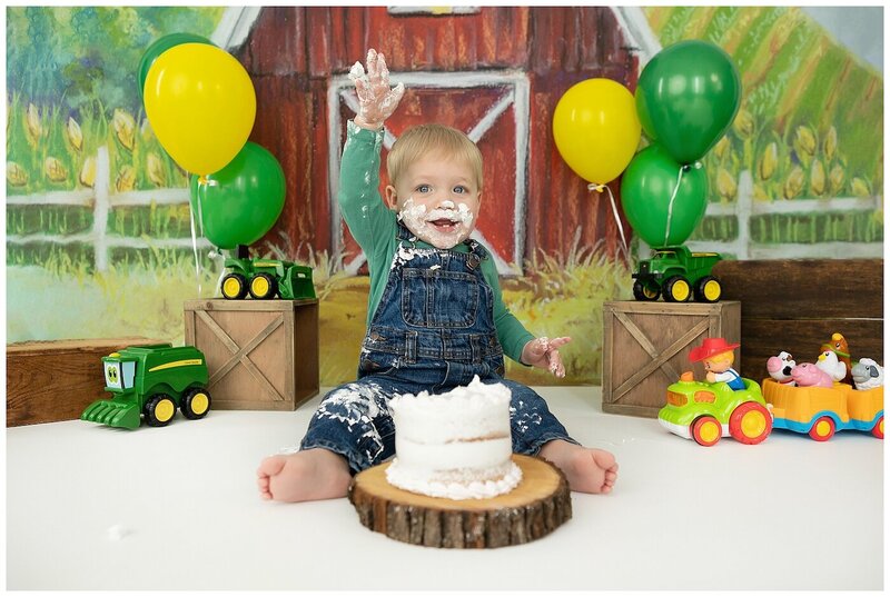 one year old boy in overalls with cake on his face surrounded by john deere toys