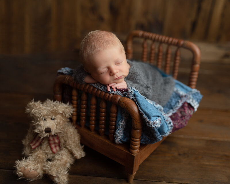Newborn boy posed on a wooden spindle bed with vintage quilt and grey sleeper beside a plush bear – a rustic and heartfelt newborn portrait.