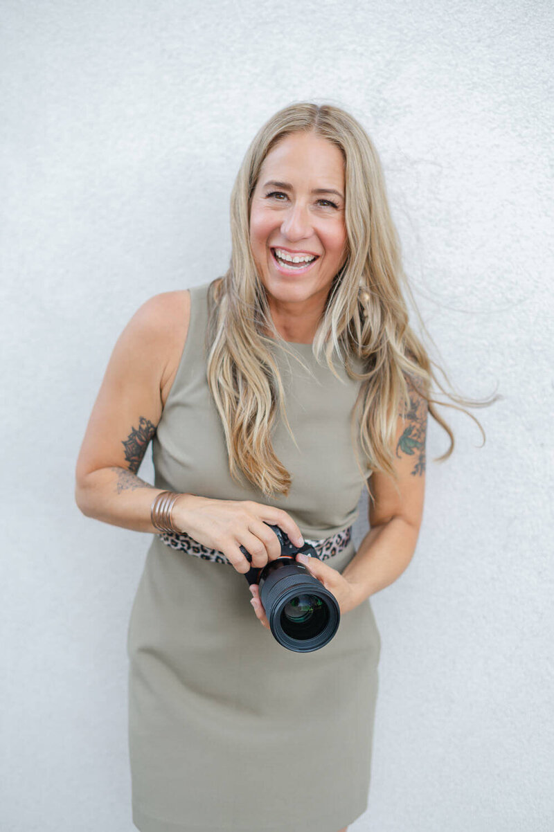 Kristen, Kelowna brand photographer wearing green dress holding camera in hands smiling at camera.