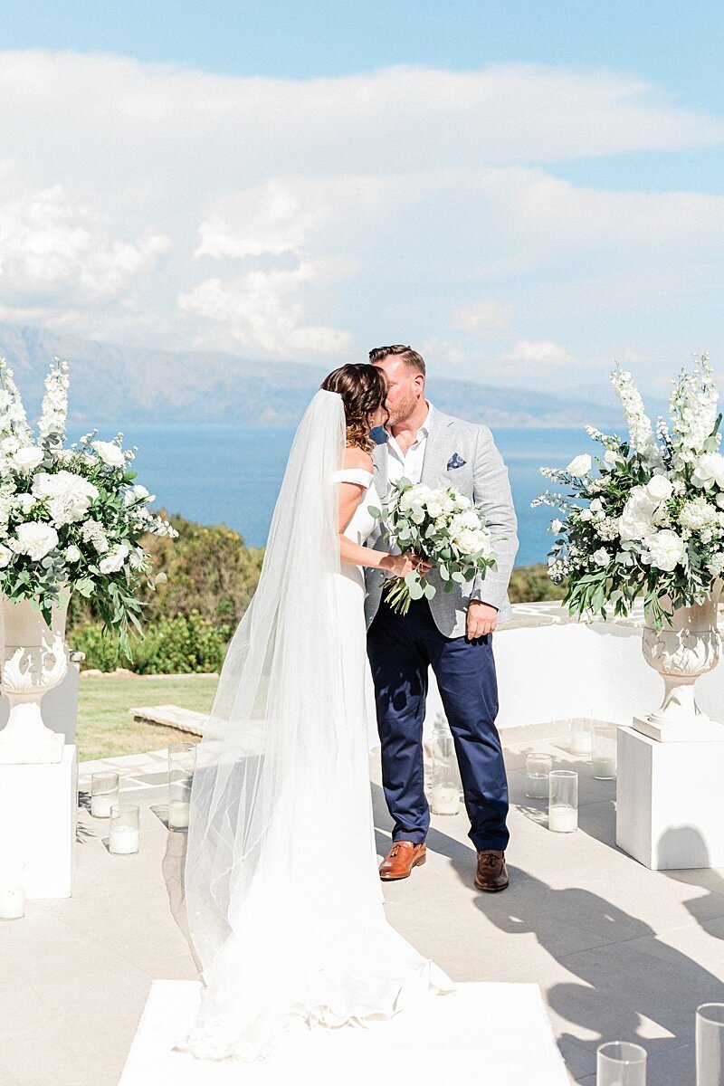 Bride and groom share a kiss at a ceremony altar framed by white florals and scenic coastal views in the background of their destination wedding