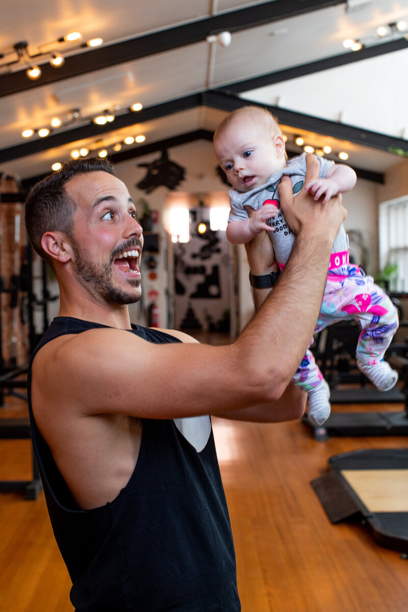 Trainer Holding a Baby in dog friendly Melbourne Gym: A smiling gym trainer joyfully lifting a baby in a family-friendly Melbourne fitness space with warm lighting and inviting decor.