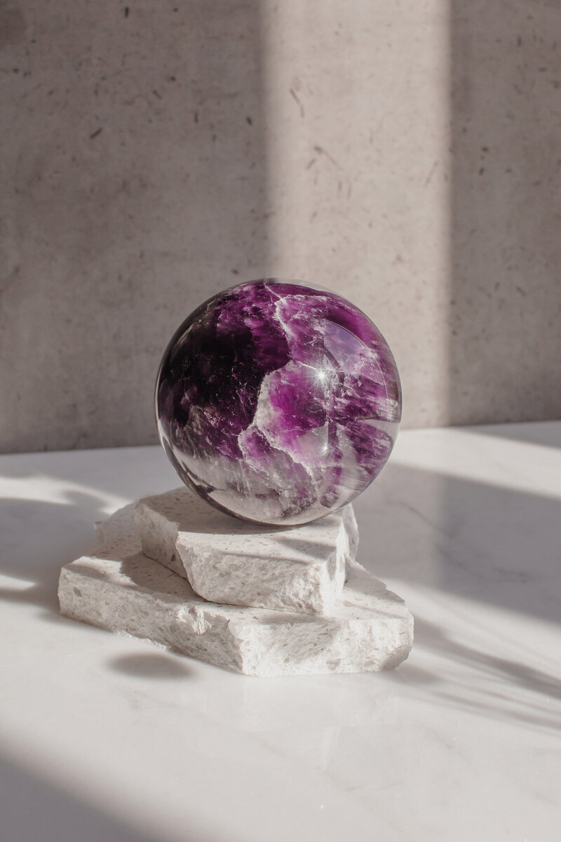 A large amethyst crystal sphere in a deep purple colour resting on quartz slabs, bathed in dappled light.