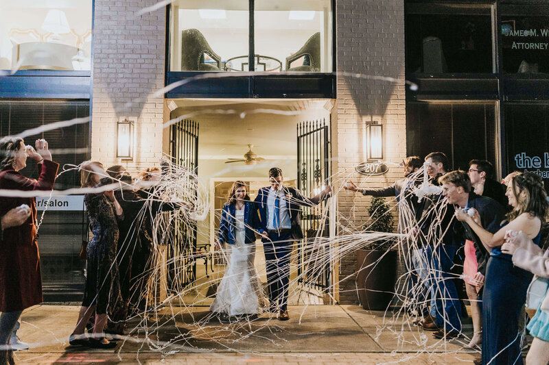 A bride and groom walk out of a building at night, smiling as guests on both sides throw white streamers in celebration. The scene is festive, with people cheering and taking photos.