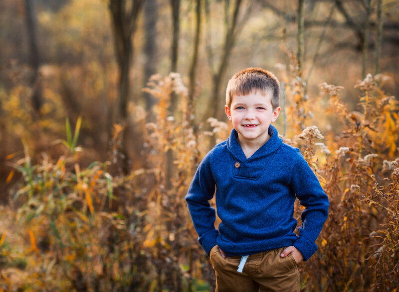 A young boy smiling outdoors in a blue sweater, surrounded by autumn foliage by Leslie Q Photography