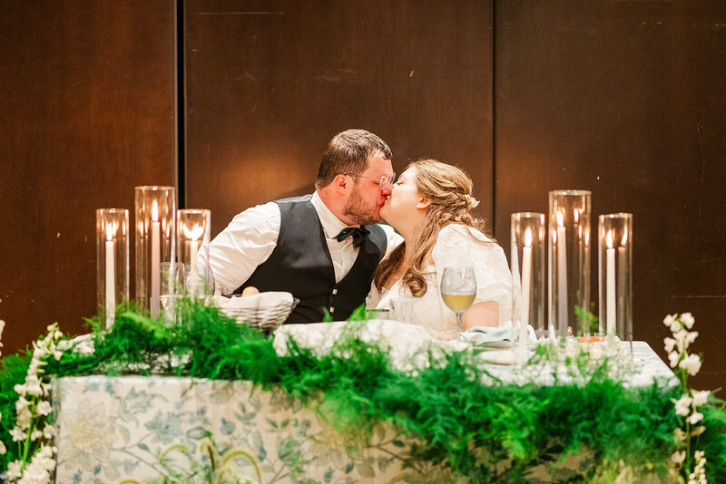 Scottish groom kissing his jewish bride while sitting at their sweetheart table in the ballroom at District Winery in DC 