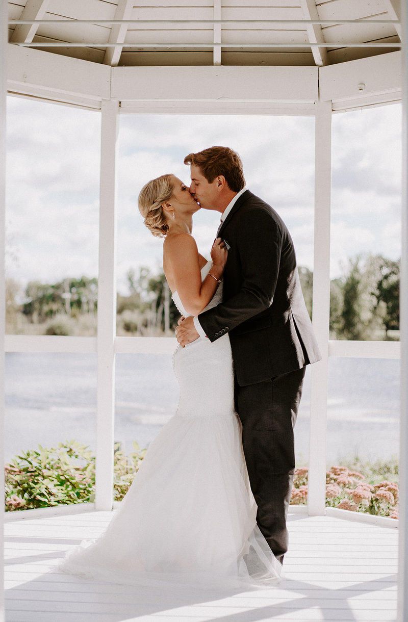 Bride and Groom kissing in a white gazebo next to a lake on their wedding day