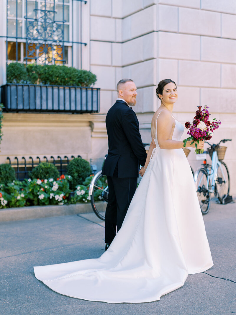 A radiant bride looking back with a joyful smile, holding a bouquet of rich burgundy and purple blooms, with her groom standing proudly beside her in a cityscape setting in front of the Hay Adams Hotel in DC.