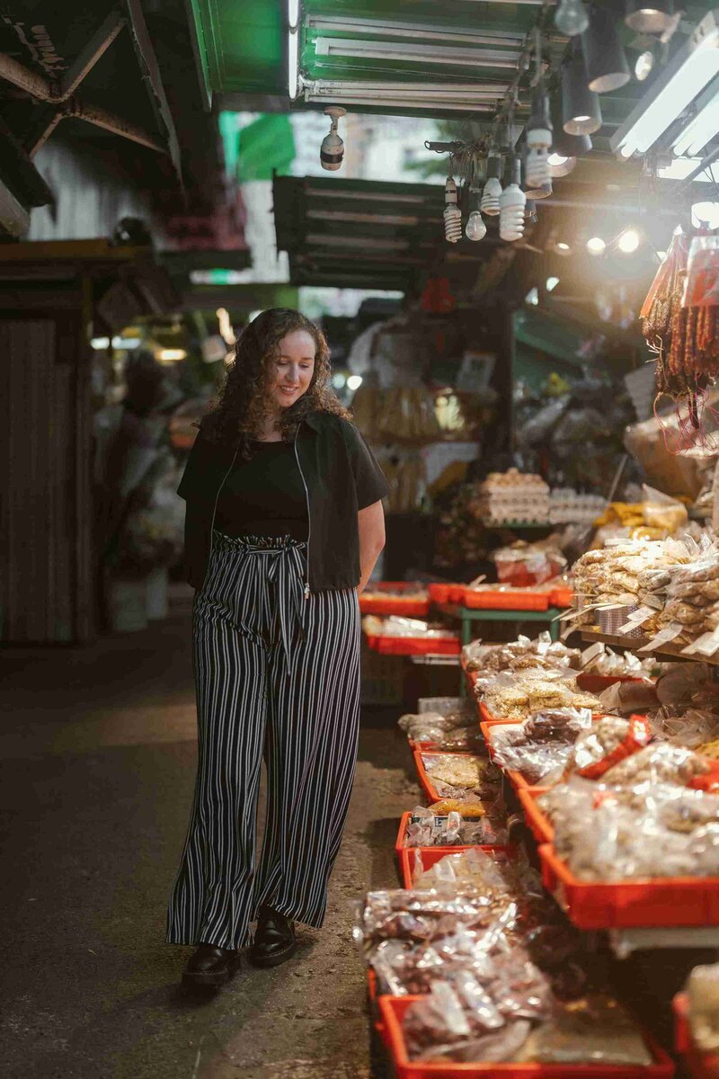 Eat Beyond Labels Owner, Christy, in striped pants strolls through a bustling, dimly lit market. She appears relaxed as she browses various packaged goods on display.