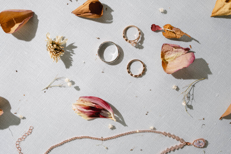 Flat lay of wedding rings, dried flowers, pink petals, and jewelry on light fabric.