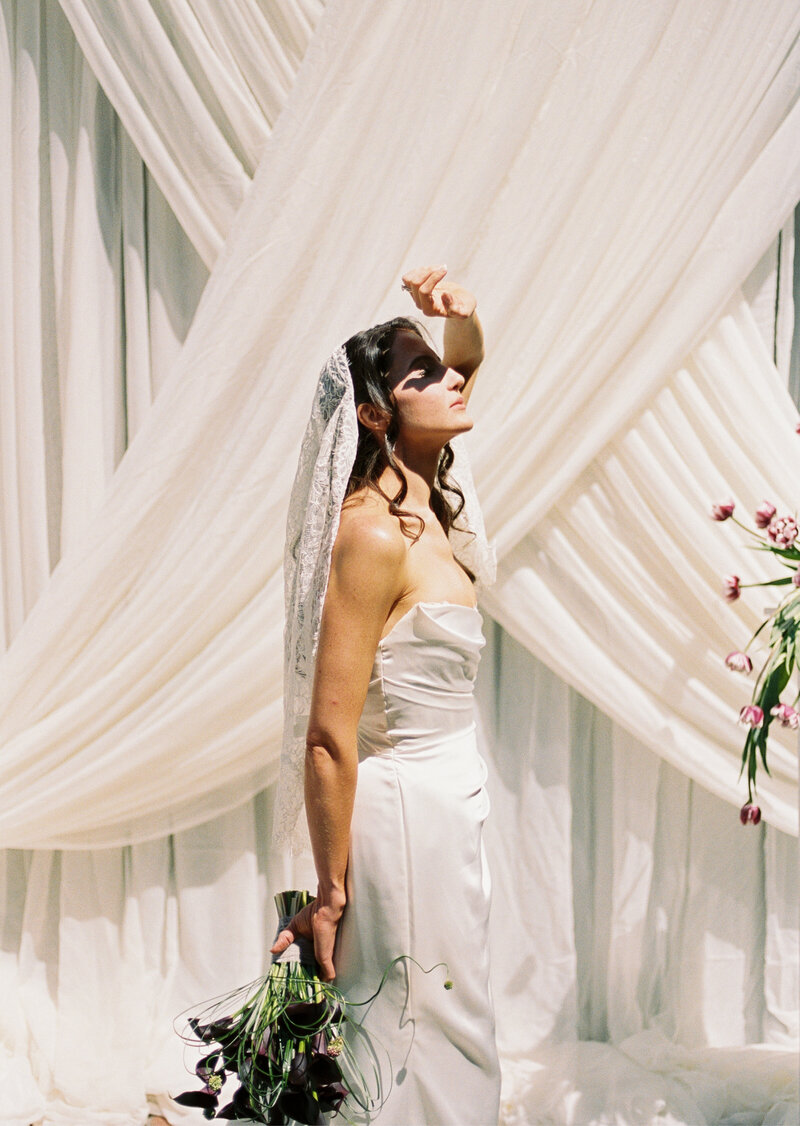 a bride standing in the sunlight blocking the sun with her hand and holding the bridal bouquet with the other hand, while standing in front of the ceremony drapery
