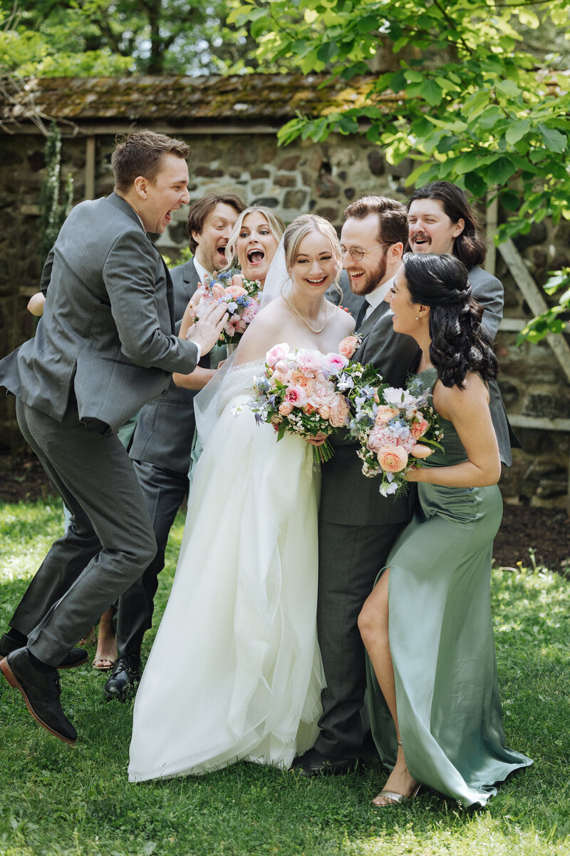Joyful wedding party photo featuring the bride and groom surrounded by their laughing bridal party in an outdoor garden setting at Parque Ridley Creek. The bride holds a vibrant bouquet of pink and pastel flowers, wearing a flowing white gown, while the bridesmaids and groomsmen in matching green and gray outfits celebrate together under lush greenery.