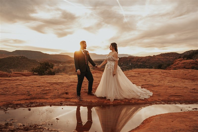 A couple stands hand in hand on a red rock formation in Sedona, Arizona, during their elopement. The setting sun casts a golden light, reflecting off a small pool of water, as they share an intimate moment against the stunning desert backdrop.