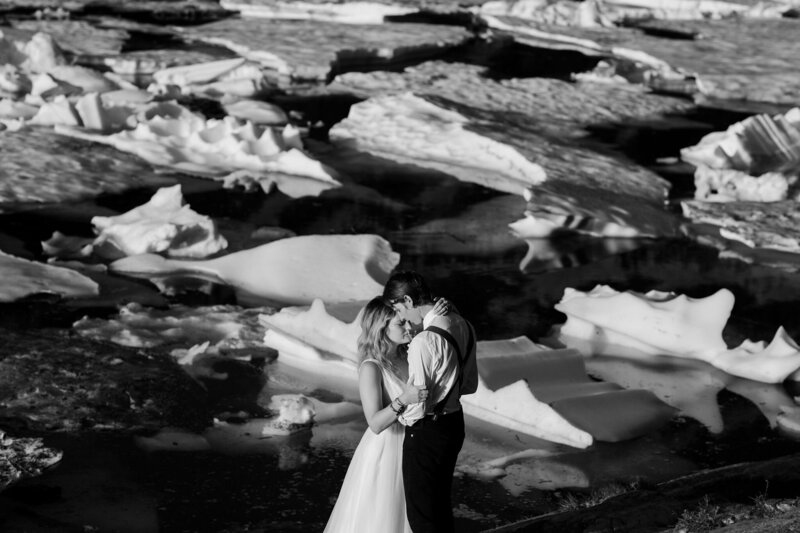 two adventure elopement photographers in their wedding attire embrace in front of a partially frozen alpine lake. chunks of small ice burgs float behind them in the background