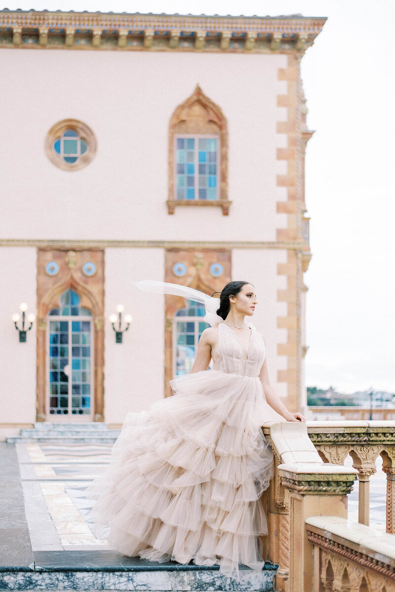 bride in a pinkish tan wedding gown standing on a staircase at the Ringling Museum as she looks out off of the balcony