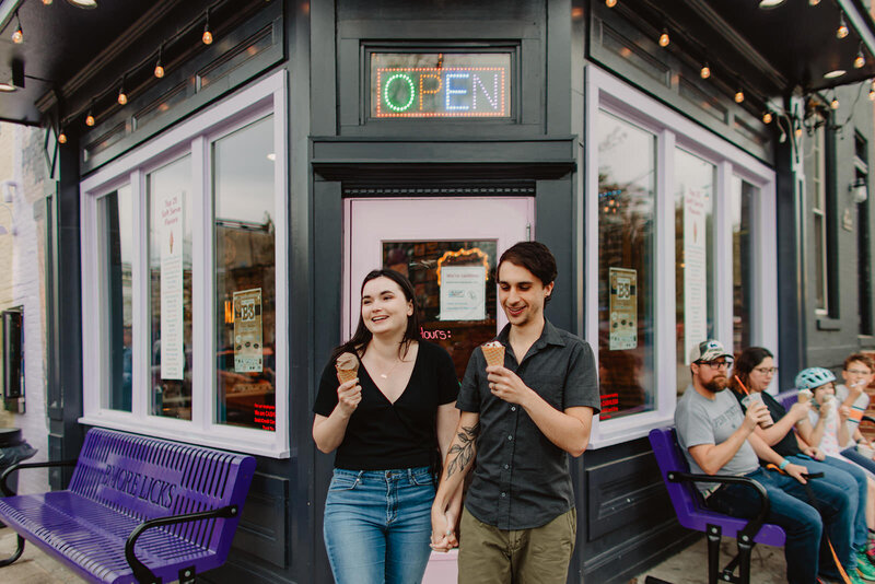 A couple holding hands and walking out of an ice cream shop with cones in each of their hands 