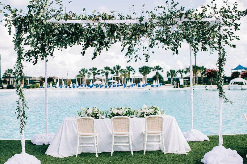 Rectangular table dressed in white linen facing a lake, with white chairs and floral centerpieces arranged beneath a greenery arch.