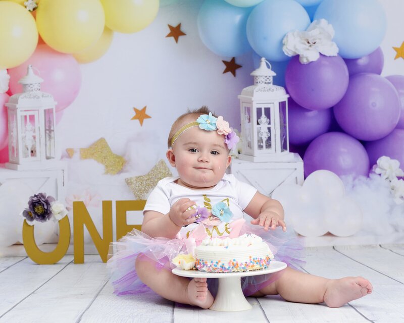 A baby girl wearing a unicorn-themed outfit with a colorful flower headband, sitting with a small cake surrounded by pastel balloons and gold stars.