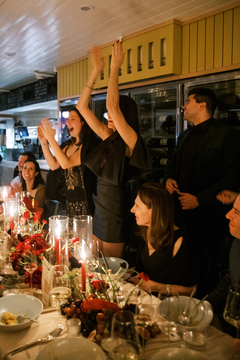 Guests celebrating at a birthday reception, raising their hands in joy, surrounded by candlelit tables with rich red floral decor.