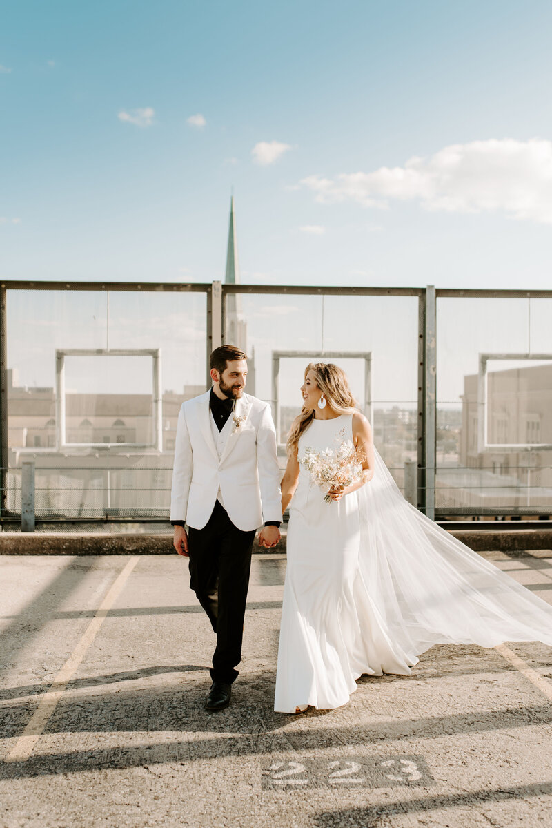 A couple walking in wedding attire on a Tulsa rooftop, holding an Anthousai bouquet.