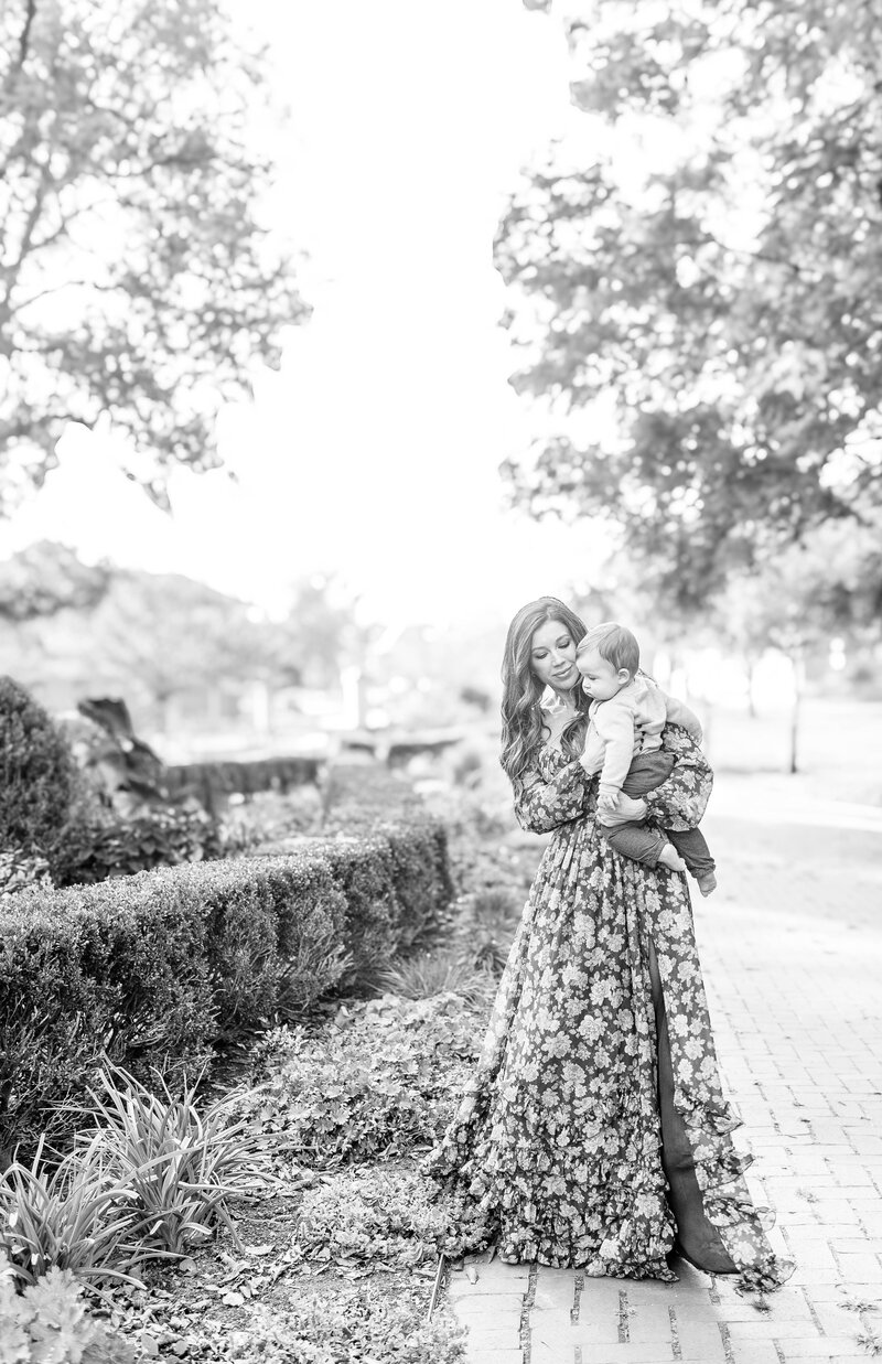 Black and white photo of a mother in a floral dress lovingly holding her toddler on a garden-lined path in a Kansas City park. 