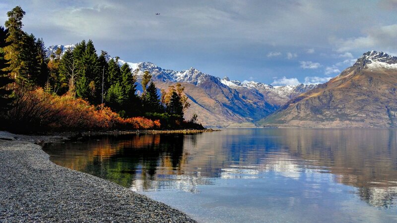 Scenic view of a calm Lake Whakatipu in Queenstown reflecting snow-capped mountains, autumn-colored trees, and a partly cloudy sky, with a rocky shoreline in the foreground