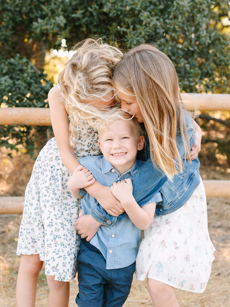 Three children embrace joyfully outdoors family session. Two girls in floral dresses hug a smiling boy in a denim shirt. Sunlit leaves create a warm, cheerful atmosphere.