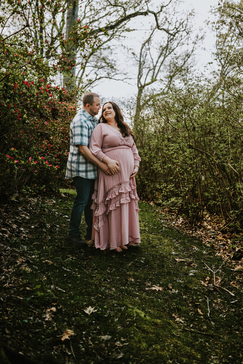 Pregnant mom in pink dress holding her belly and leaning back on husband while looking lovingly at him in garden in Montgomery County Maryland photographed by Bethany Simms Photography