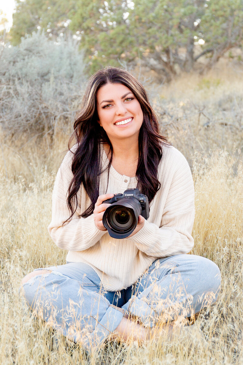 A young woman sits amidst the sagebrush and trees in a desert landscape while holding her camera and smiling at the camera.