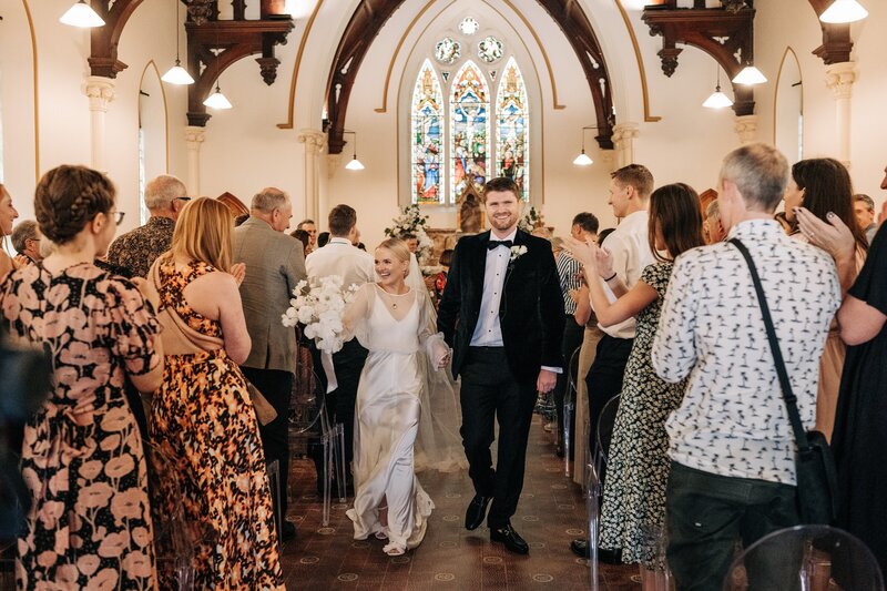 a couple walk down the aisle in an old chapel in christchurch after their wedding