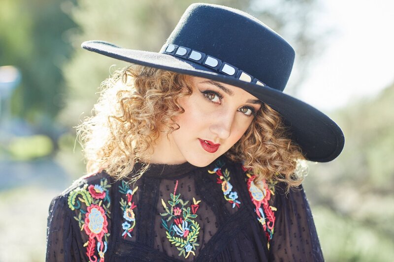 Whimsical teen model with tight curls, bold makeup, and a wide-brim hat wearing an embroidered sheer top – styled during a workshop session.