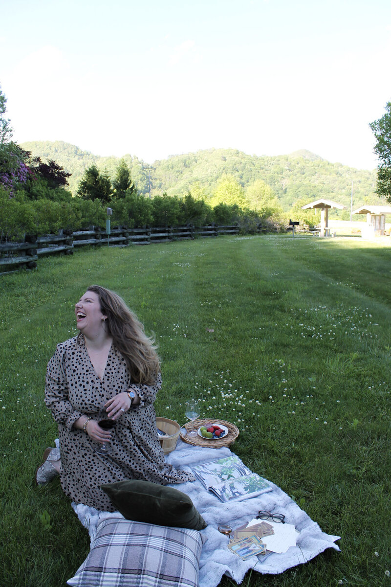 Image of Ashleigh Henry in a field with mountains in the background laughs at a subject off camera while holding red wine, reflecting on her work with emotionally intelligent women ready to reorient their lives with clarity and reverence.