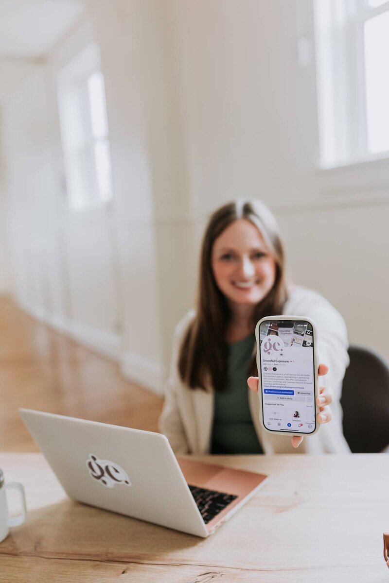 McKenna Fahrenbruch, social media strategist at Graceful Exposure Media, sitting at her desk with her laptop and holding her phone showing a Facebook page—highlighting expertise in social media content, community growth, and engagement."