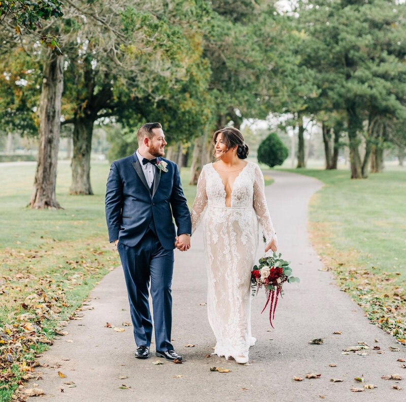 A bride and groom walk hand-in-hand down a tree-lined path on their wedding day. The bride wears a long-sleeved lace dress  and holds a bouquet of flowers, while the groom is in a blue suit with a bow tie. Both are smiling at each other