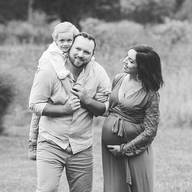 Dad, son and expecting mother walk through field smiling and laughing with each other.