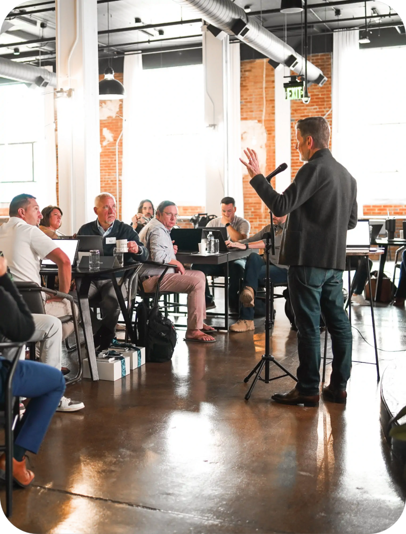 Michael Hyatt addressing a seated group in a bright industrial-style room, standing at a microphone near the front.