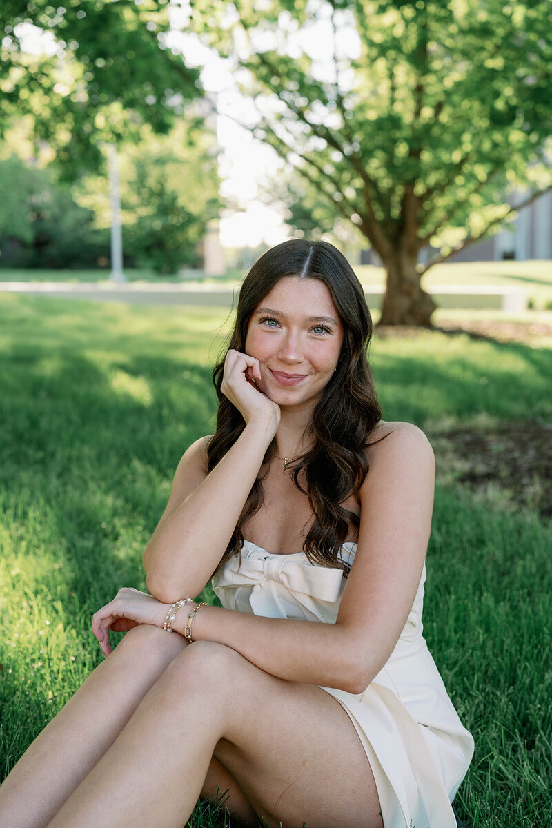 Senior woman sitting in tall grass and smiling at the camera during her WMU graduation photos.