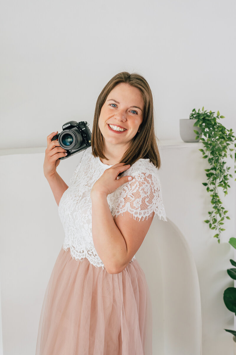 denver photographers headshot with photographer holding her camera while wearing a white loace top and smiling at the camera