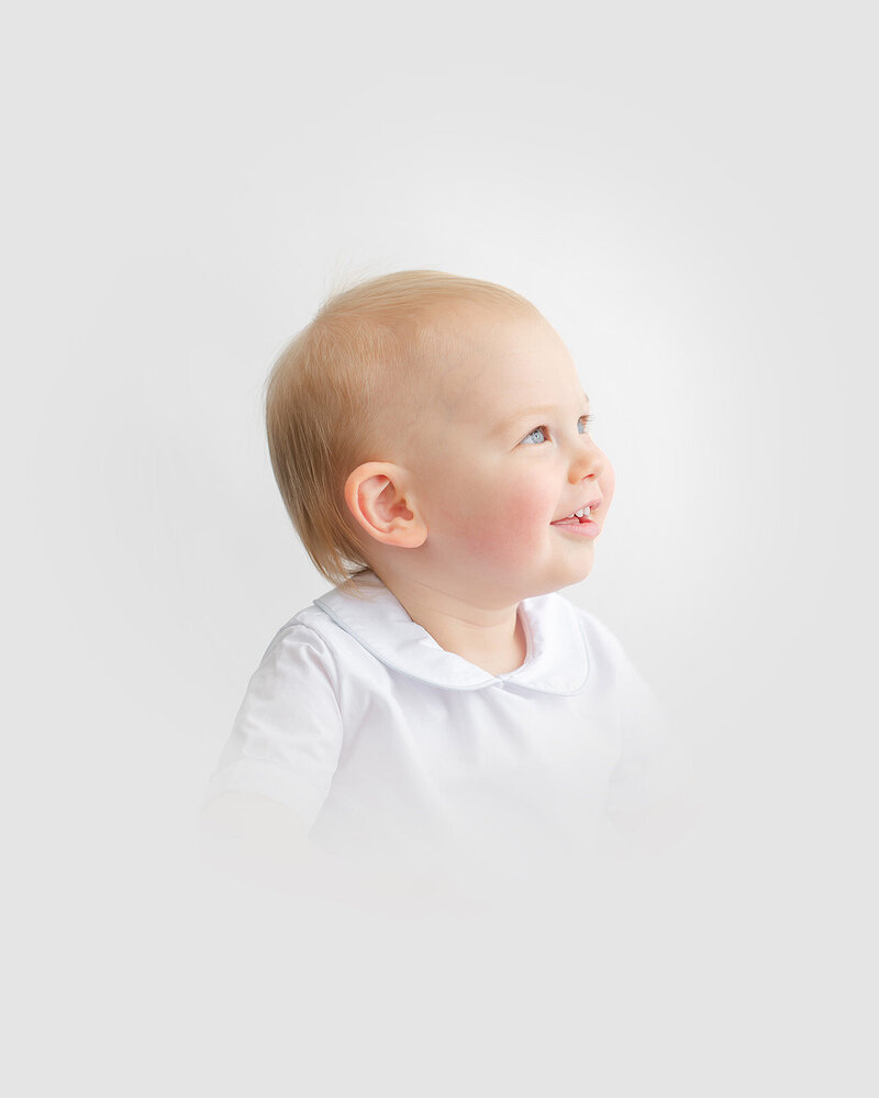A baby with light hair and a faint smile looks to the side, wearing a white shirt with a collar against a timeless white background.