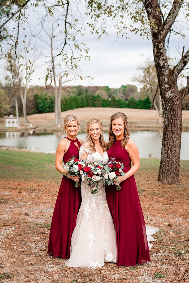 Bride with her two bridesmaids standing on her sides wearing red with a pond in the background