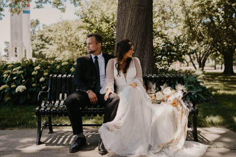 bride and groom sit on park bench looking in opposite directions after just getting married while still in their wedding attire. Bride wears a beaded pearl wedding dress with large bouquet placed beside her.