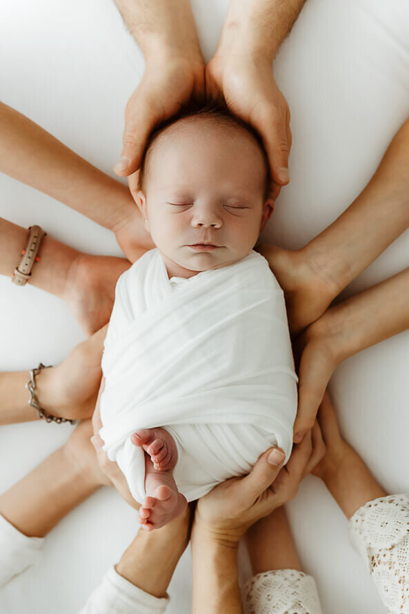 Baby boy sleeps while family all has hands touching him.
