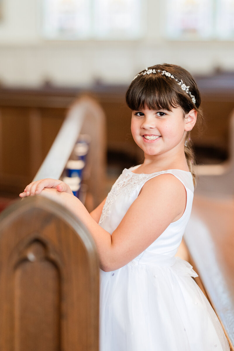 Young girl in white dress posed in a pew in a church.
