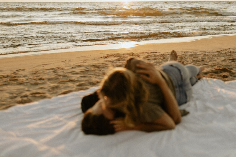 A romantic photo of a couple on a white blanket at the beach overlooking Lake Michigan during sunset. 