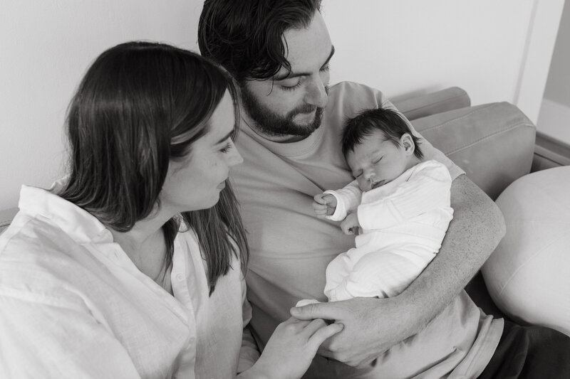 A heartwarming black and white portrait of new parents in Portland, gazing lovingly at their newborn cradled in the father’s arms. This intimate moment captures the pure joy and tender emotions of early parenthood