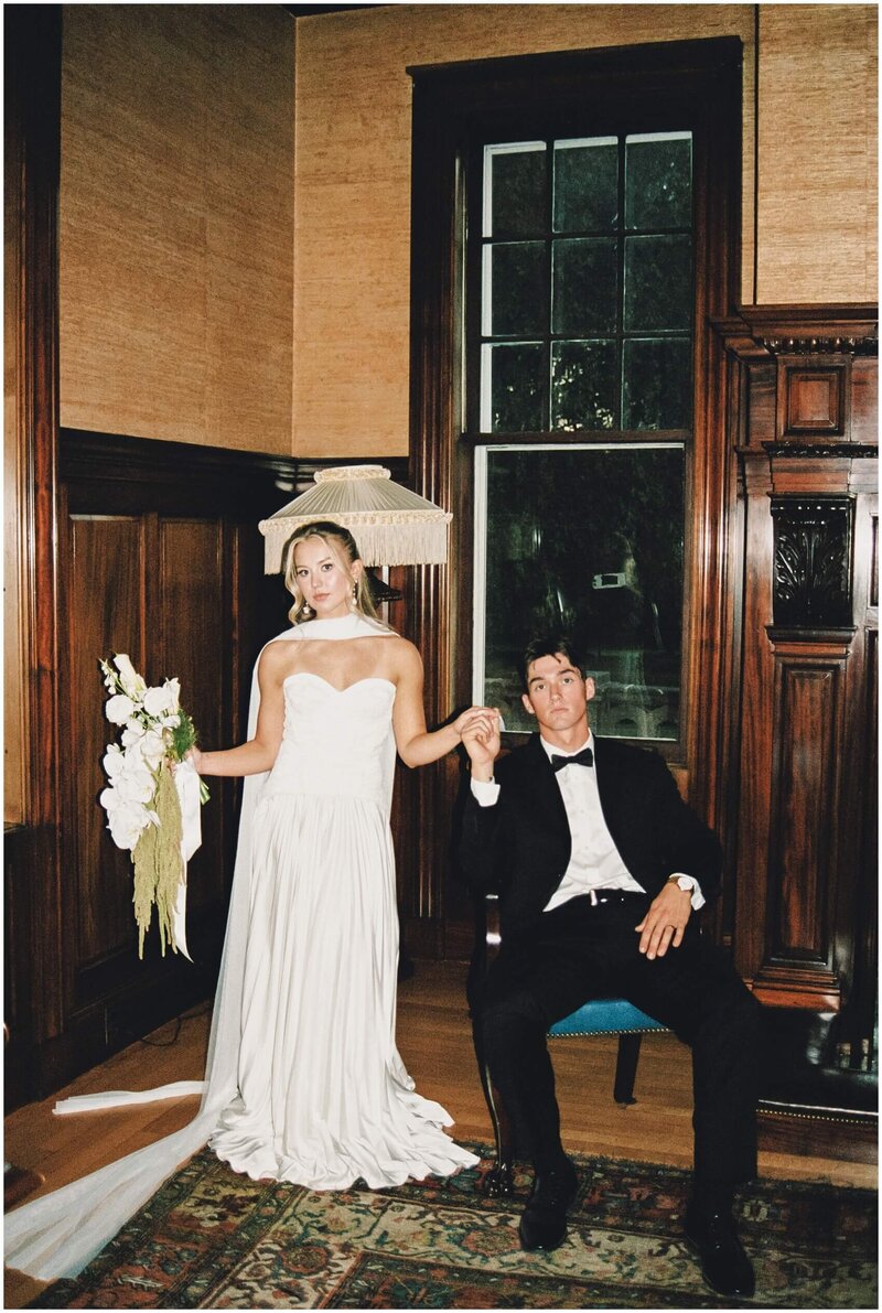 A groom in a black tuxedo sits in an antique chair holding the hand of his standing bride holding her white bouquet