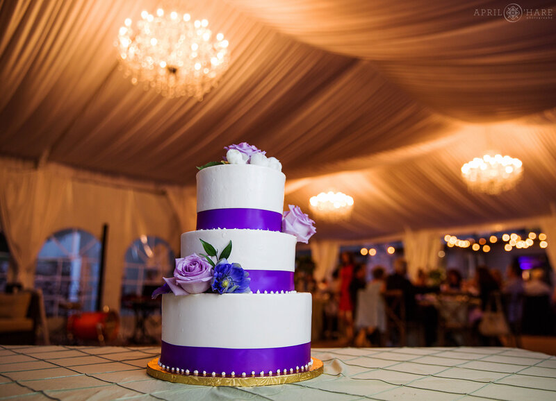 Prettywhite cake with purple ribbons and flowers decorating it with chandeliers inside a white tent hanging in the backdrop at Boulder Creek Wedgewood Weddings