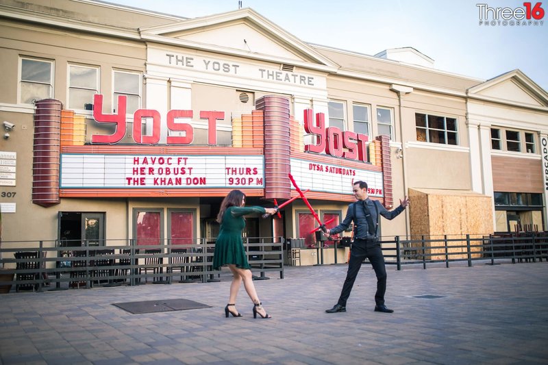 Engaged couple square off with light sabers during engagement photo shoot in front of the Yost Theater in Santa Ana