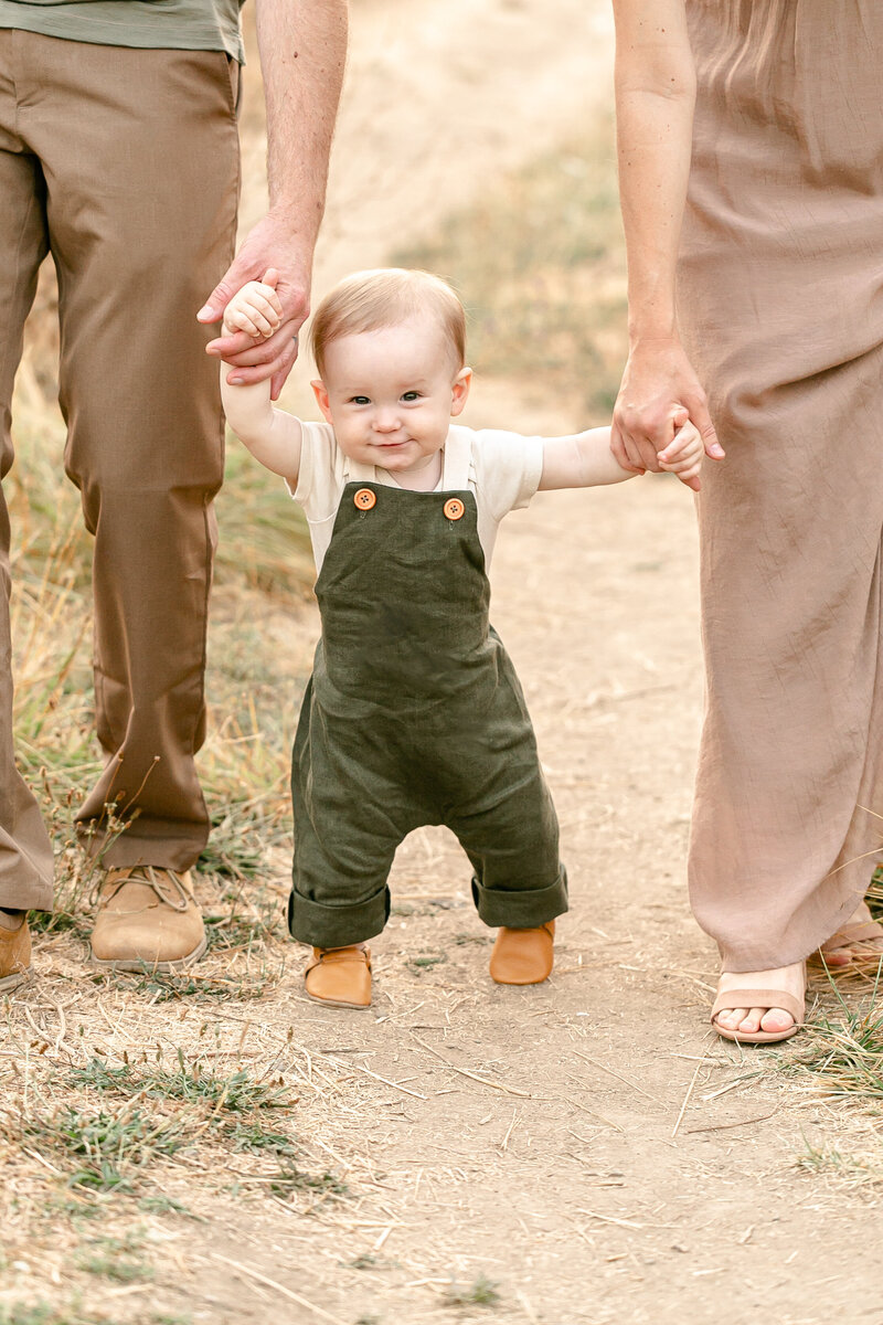 One year old baby wearing olive green overalls is walking while holding both parents hands outdoors on a dirt path. He is smiling up at the camera. Portland Portrait Photography.