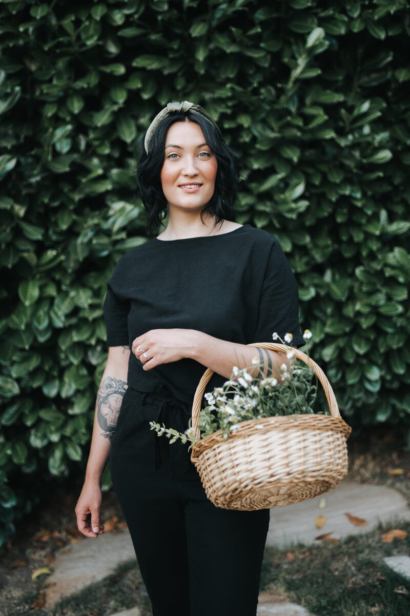 Woman wearing headband in an all-black outfit holds a woven basket full of herbs as she smiles at the camera.